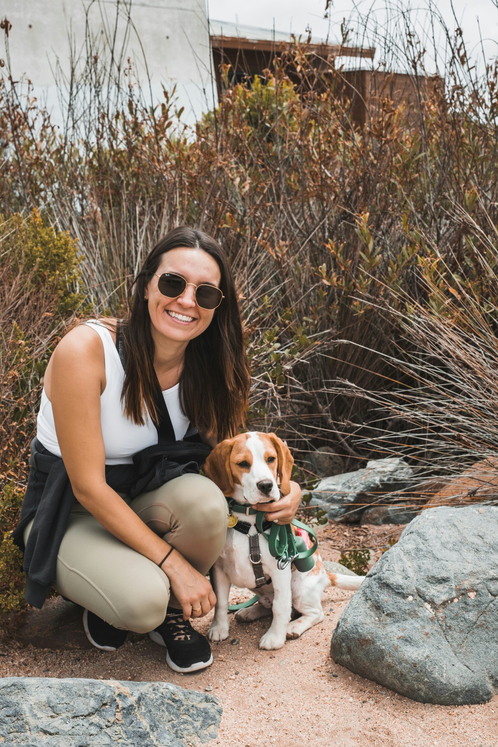 Woman in sunglasses kneeling beside a beagle dog on a nature trail.