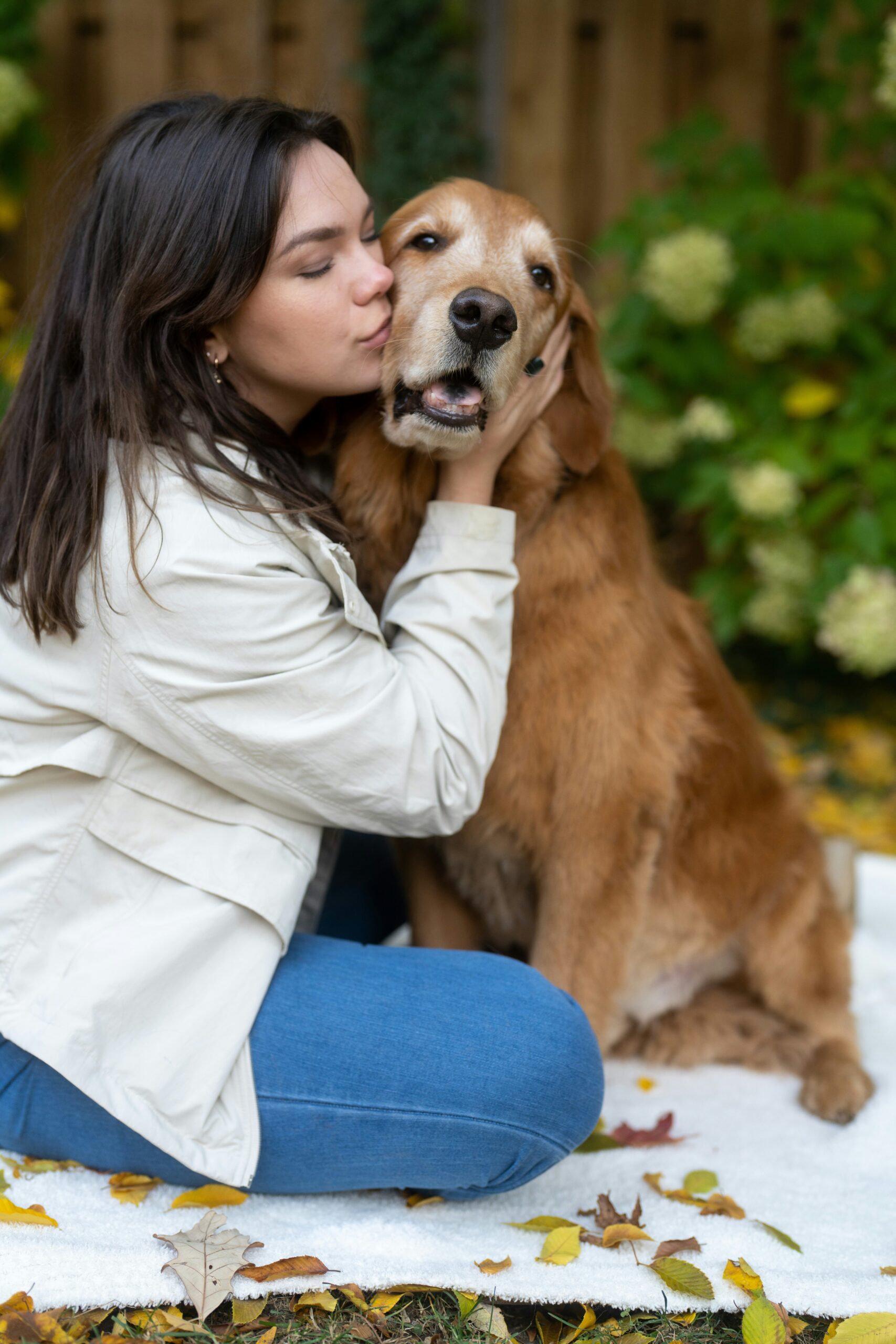 Woman kissing her Golden Retriever on a blanket in a garden with autumn leaves
