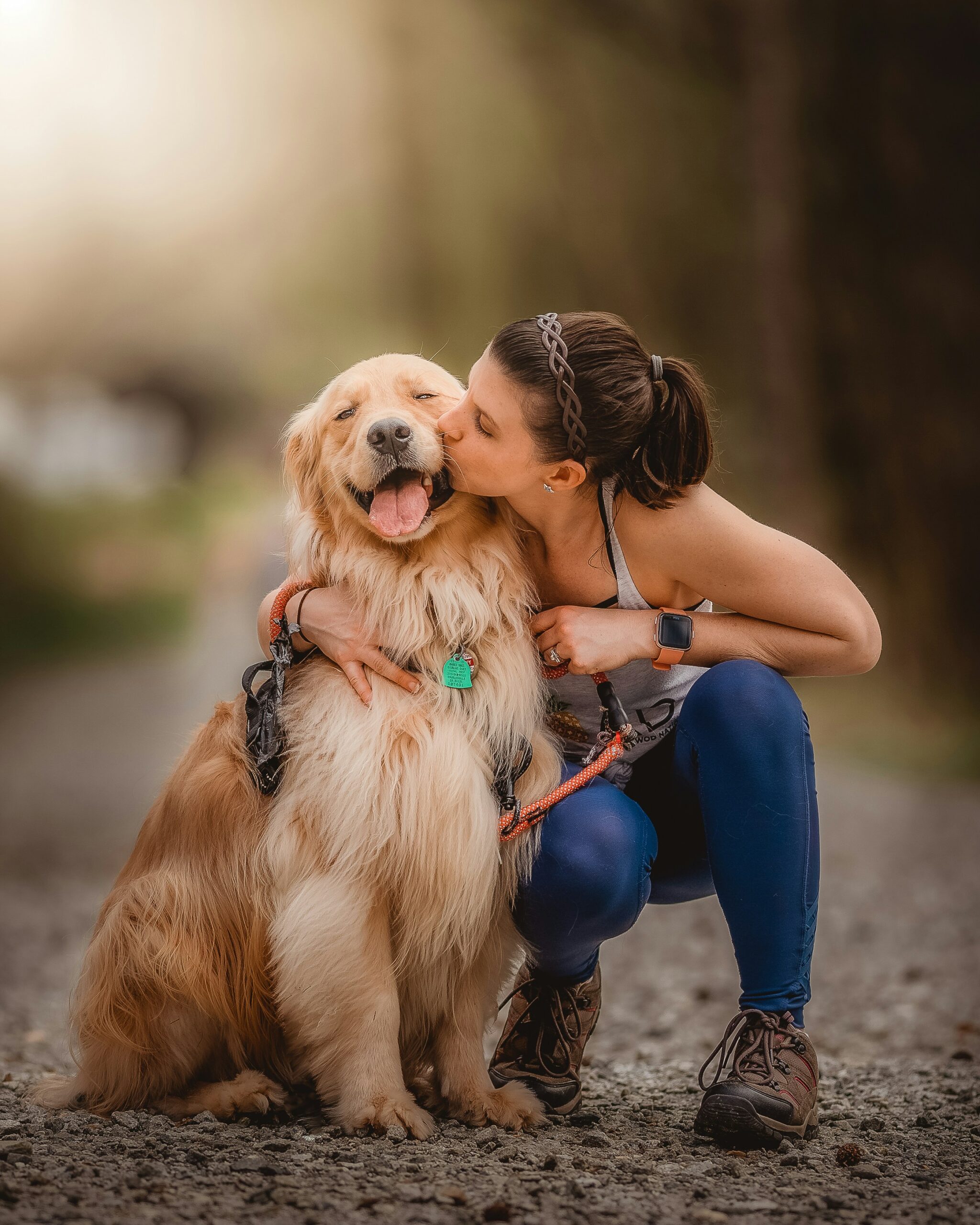 Woman hugging and kissing a happy golden retriever on a sunny outdoor trail