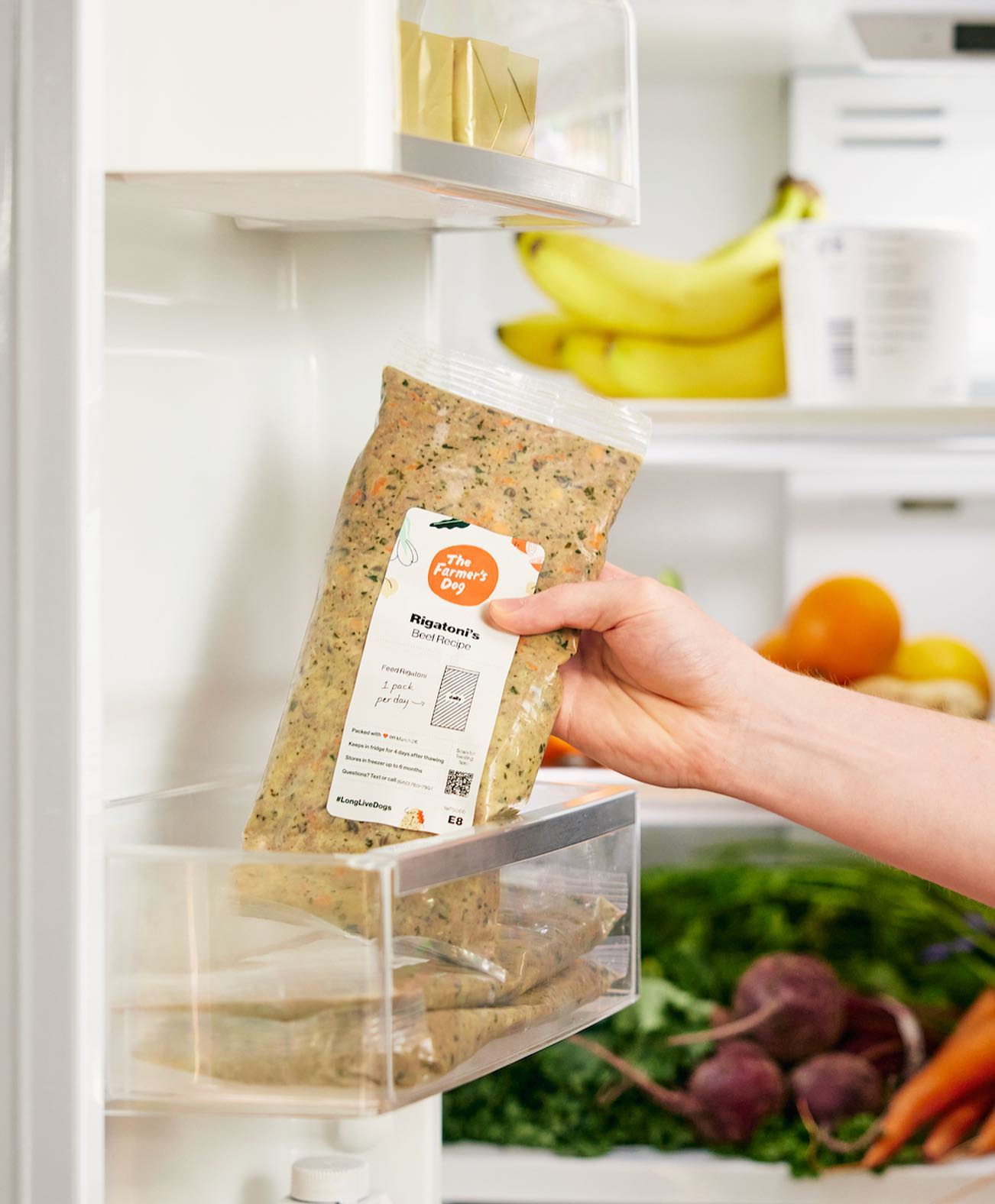 Hand placing The Farmer's Dog fresh beef recipe into refrigerator drawer with fruits and vegetables.