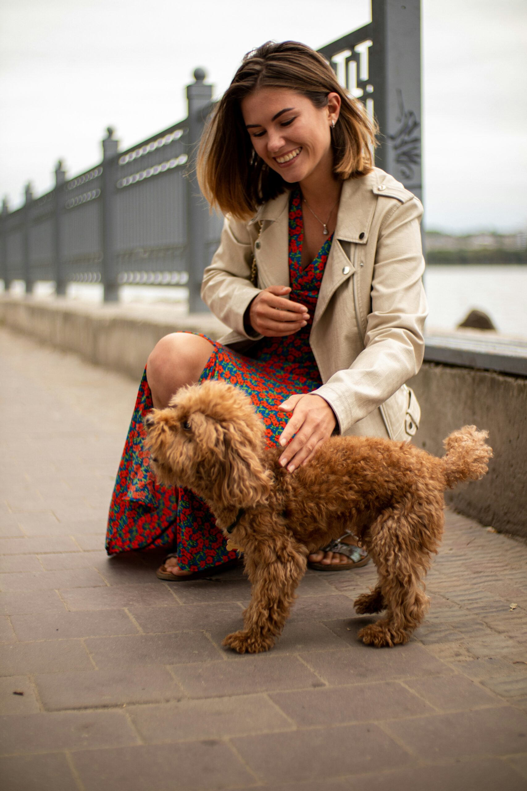 Woman in a floral dress petting a small brown poodle on a riverside promenade.
