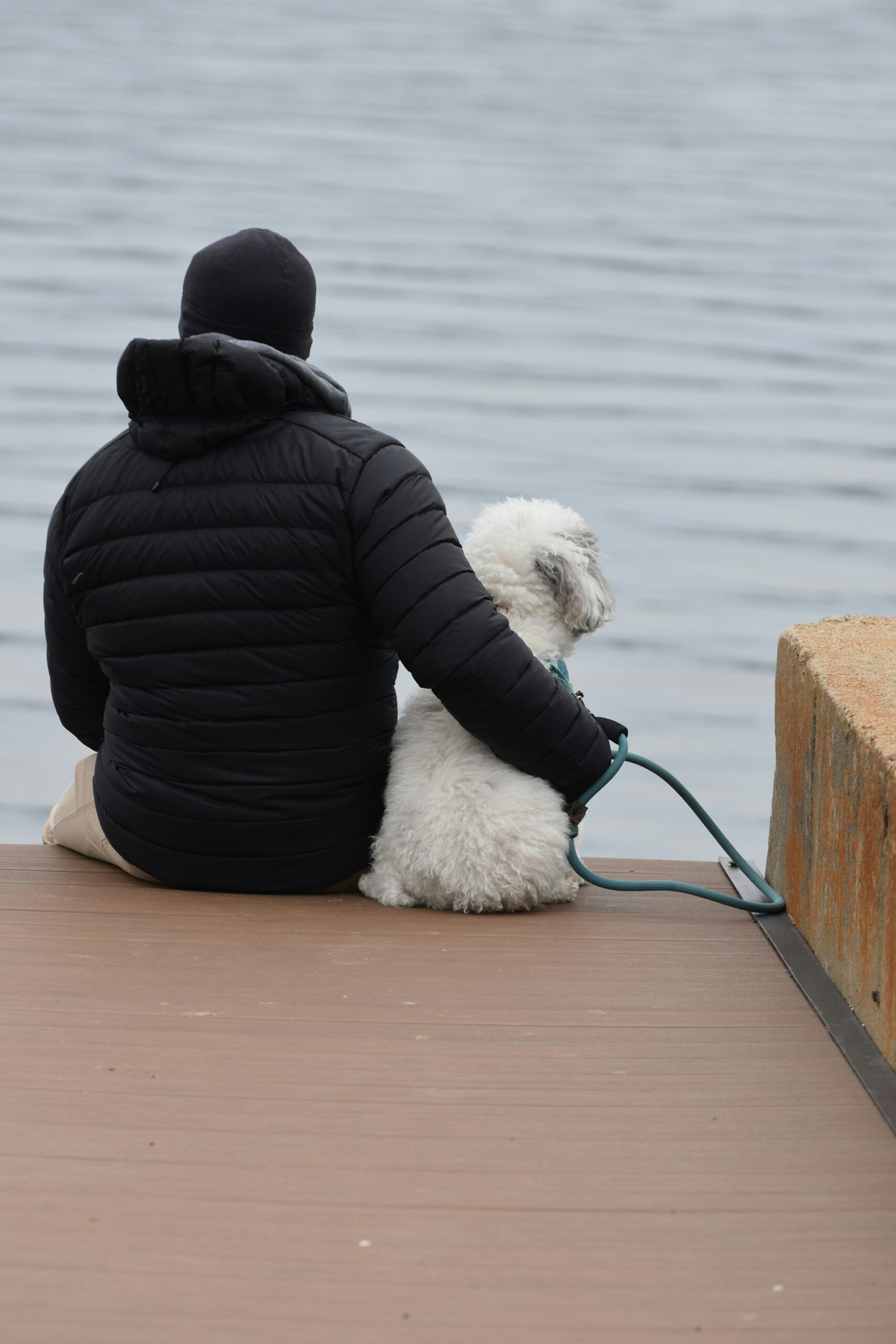 Person in black jacket sitting with a white poodle on a dock by the water.