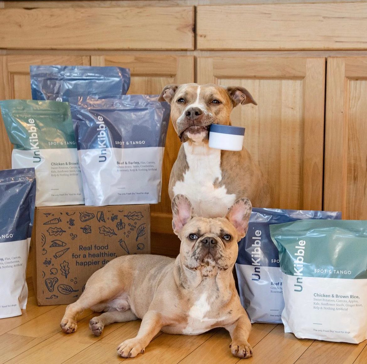 Two dogs with several bags of UnKibble by Spot & Tango, one dog holding the food scoop in its mouth