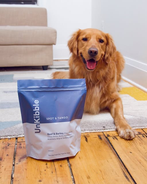Golden Retriever smiling next to a bag of Spot & Tango UnKibble Beef & Barley in a living room setting