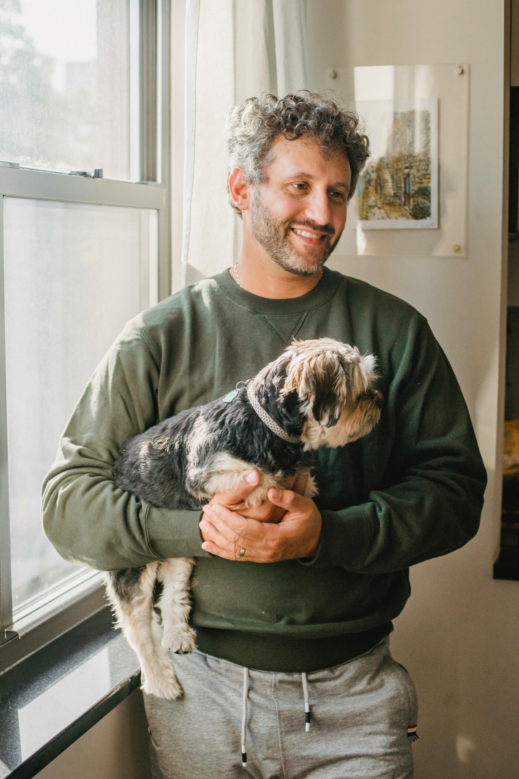 Man in a green sweatshirt holding a Yorkshire Terrier by the window, both looking out toward the light